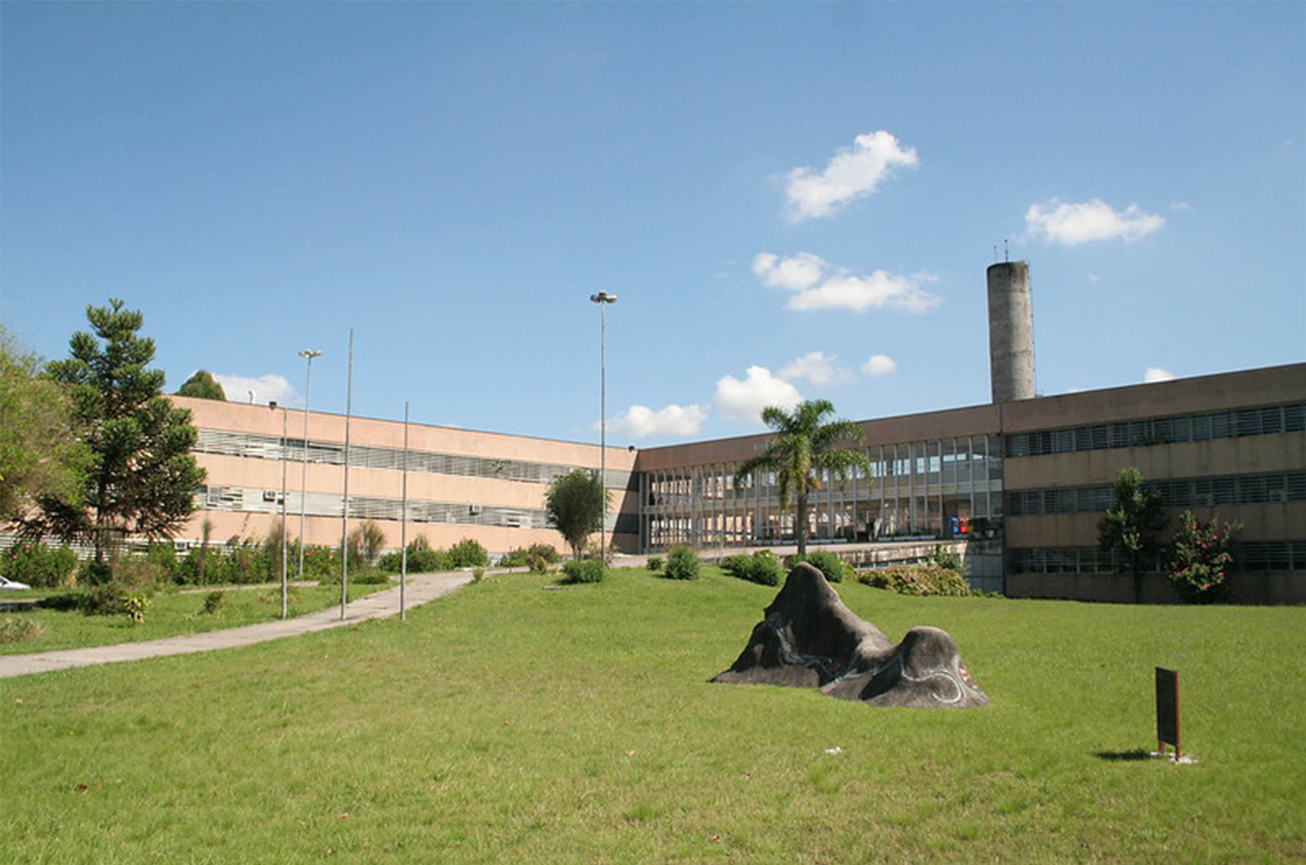 Prédio perpendicular em segundo plano, e em primeiro uma grama com duas pedras e algumas árvores.