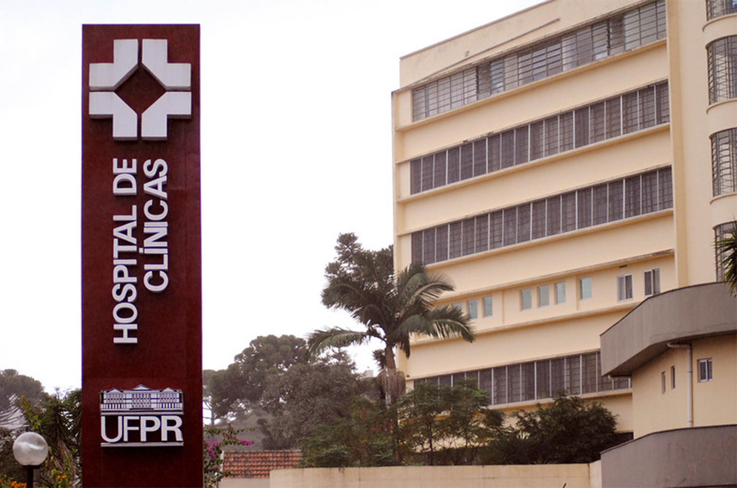 Placa escrito "Hospital de Clínicas" em primeiro plano e ao fundo um prédio de cinco andares amarelo e árvores.