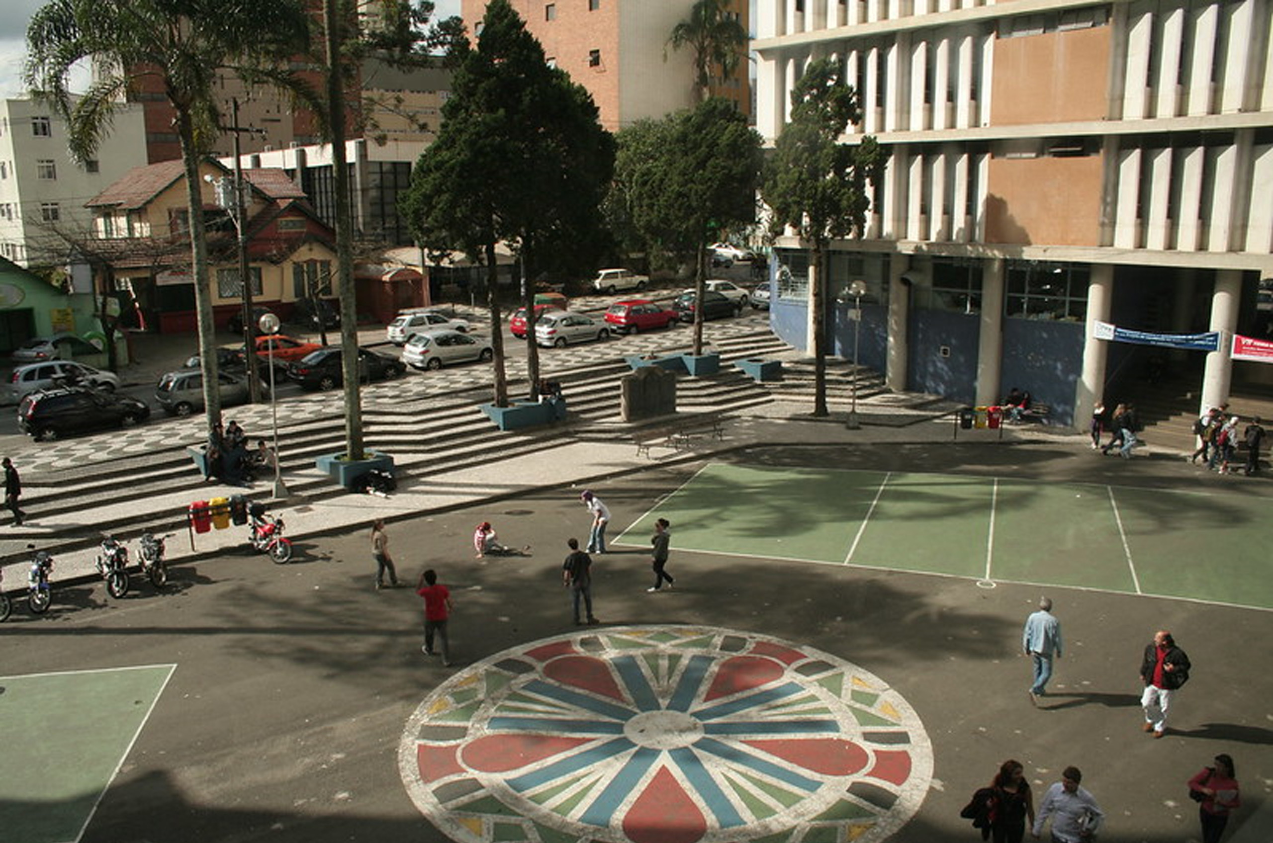 Quadra com escadas entre prédios com alunos, árvores, carros e motos, bancos e um prédio lateral com muitas janelas.