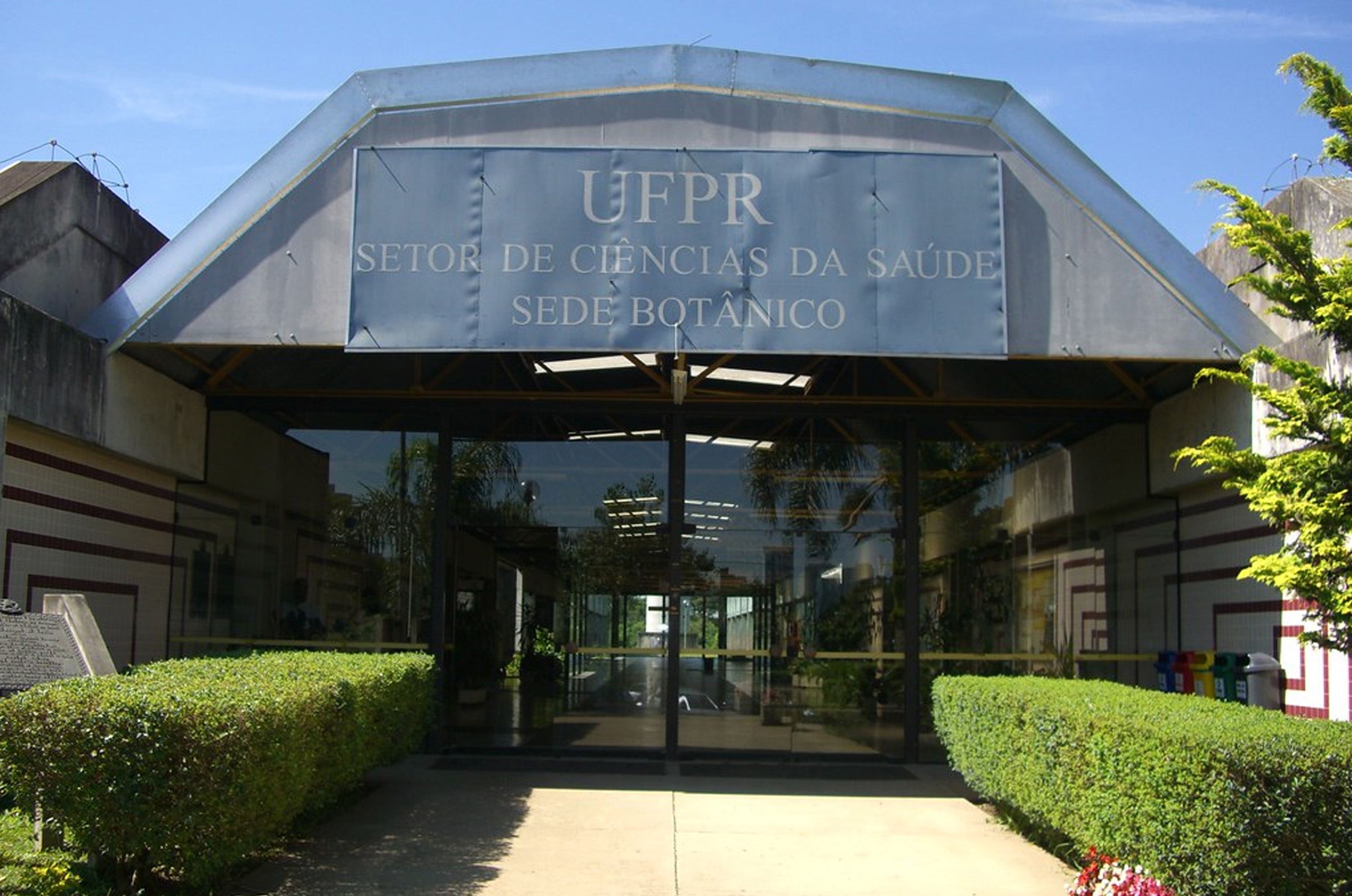 Entrada de prédio com a placa escrito "UFPR Setor de Ciências da Saúde, Sede Botânico". Com plantas em formato quadrado nas laterais da entrada.